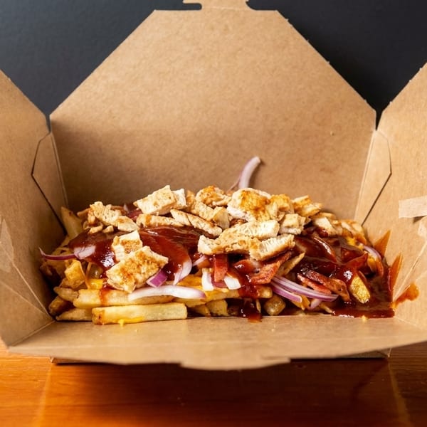 Loaded fries with chicken, bacon, red onion, and barbecue sauce in a brown takeout box.