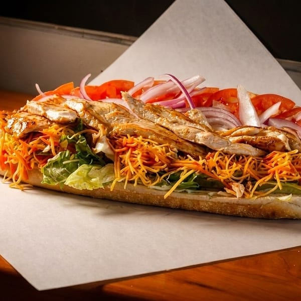 A grilled chicken sub sandwich with lettuce, shredded carrots, tomatoes, and red onions.