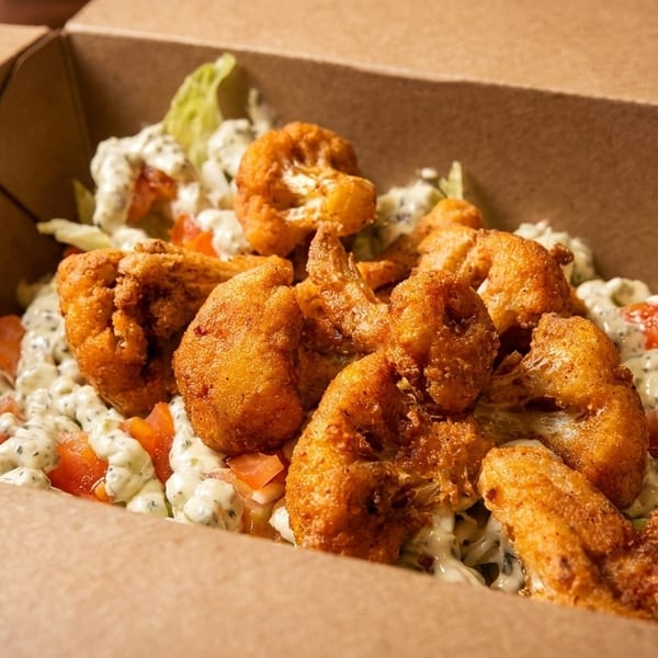 Crispy fried cauliflower bites with creamy sauce, lettuce, and diced tomatoes in a takeout box.